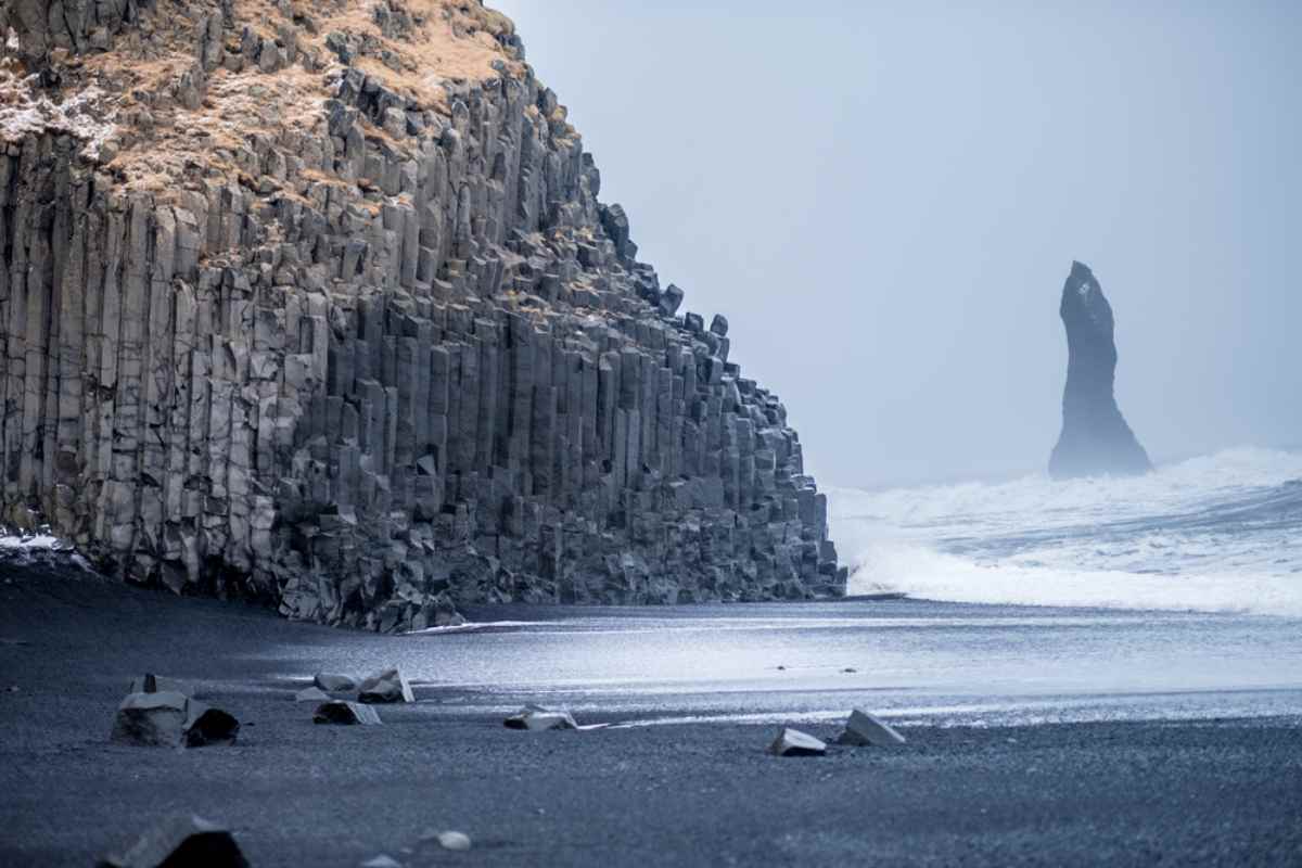 Colonne di basalto a Reynisfjara su spiaggia di sabbia nera, onde invernali e faraglione nella foschia lungo la costa sud dell&rsquo;Islanda.