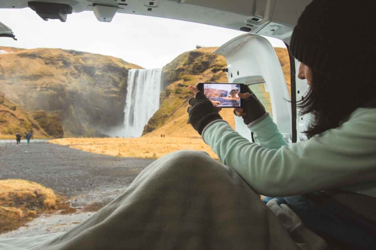 Viaggiatrice fotografa Sk&oacute;gafoss dal portellone del camper, coperta sulle gambe; giornata grigia e paesaggio dorato della costa sud.