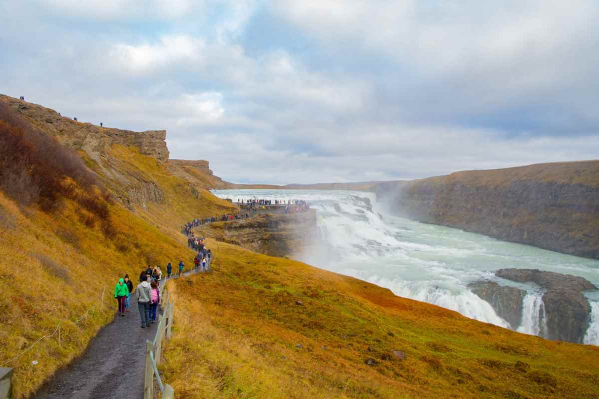 Visitatori lungo il sentiero panoramico verso la cascata Gullfoss, canyon dorato e spruzzi sotto un cielo nuvoloso nel Circolo d&rsquo;Oro.