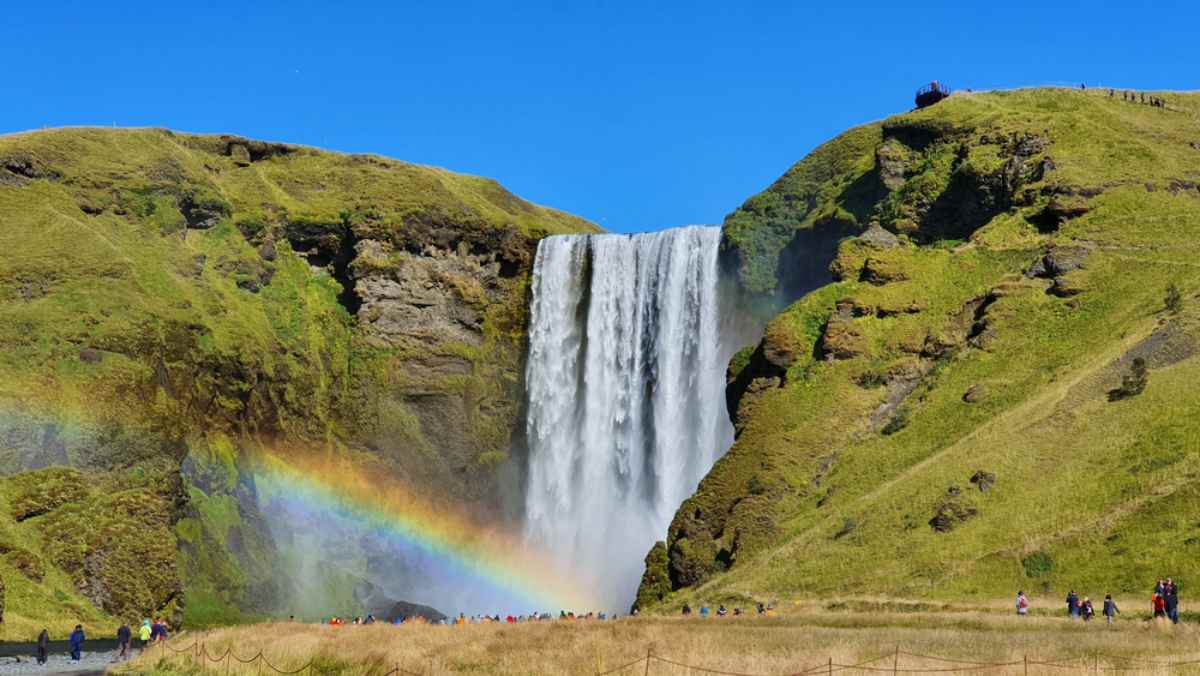 Cascata Sk&oacute;gafoss in giornata limpida con arcobaleno alla base, pendii verdi e visitatori sul sentiero di fronte allo spray.