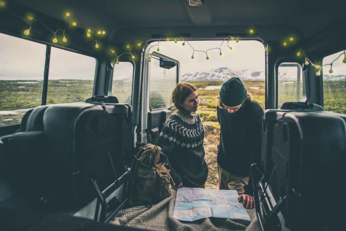 Coppia dentro un van con lucine, maglioni islandesi e mappa aperta; pianificazione dell&rsquo;itinerario invernale tra valli e cime innevate.