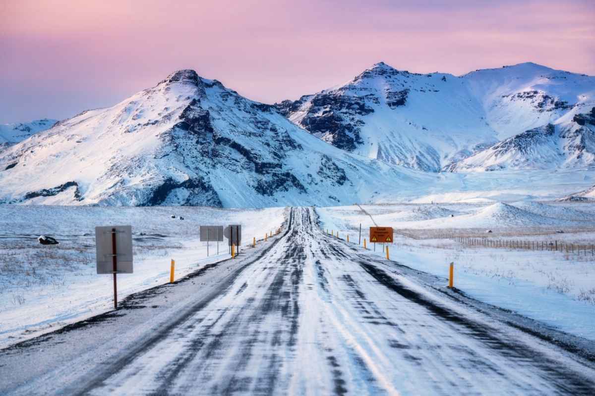 Tratto della Ring Road coperto di neve al tramonto, paletti gialli e cartelli di avviso con montagne innevate ai lati; condizioni invernali tipiche.