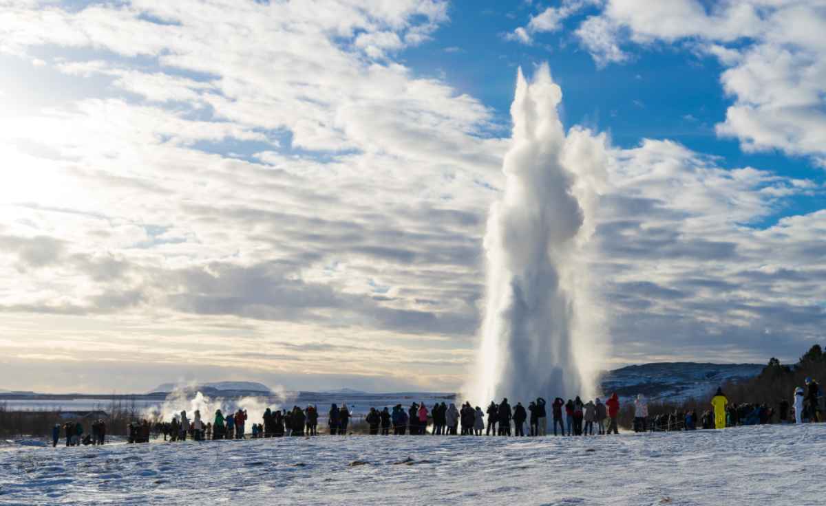 Geyser Strokkur erutta tra neve e nuvole; gruppo di visitatori osserva da lontano in una fredda giornata invernale nel Golden Circle.