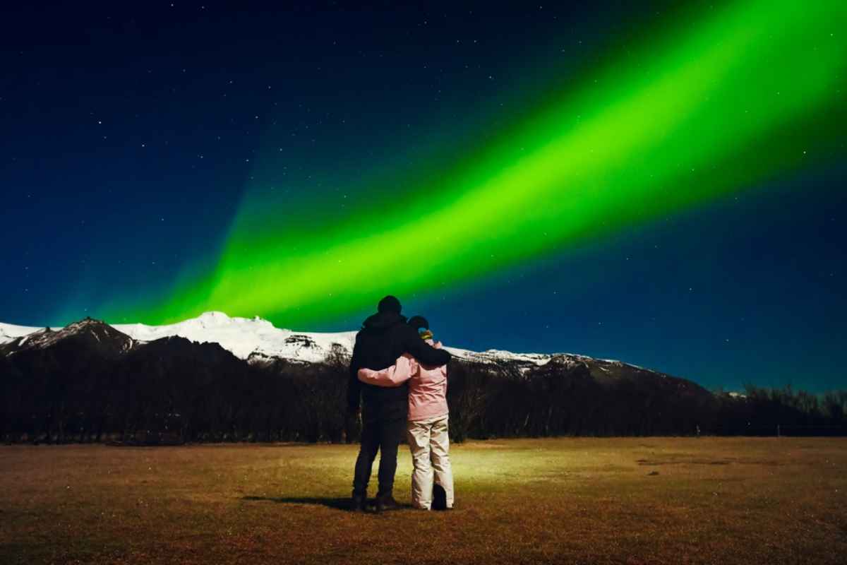 Due persone abbracciate osservano un nastro verde di aurora boreale sopra montagne innevate, cielo stellato d&rsquo;inverno in Islanda.