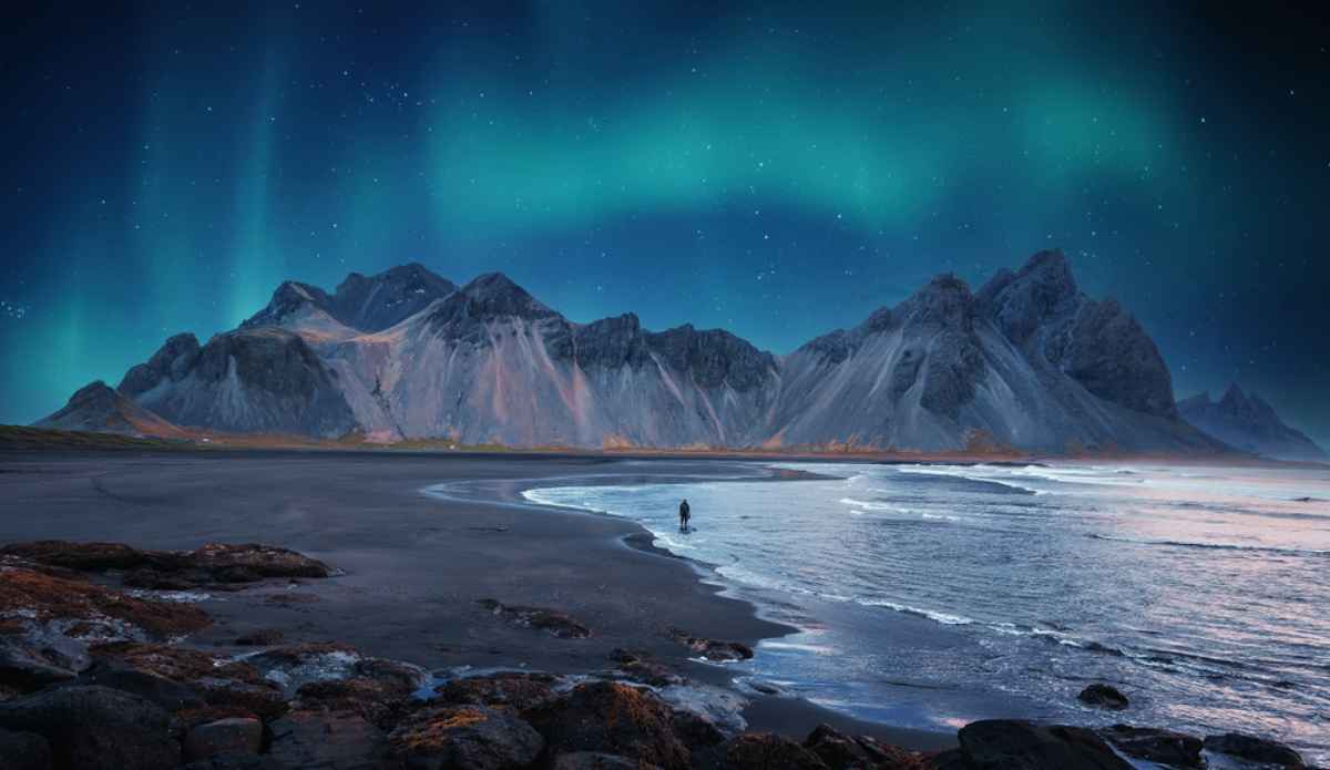 Spiaggia nera e montagne di Vestrahorn illuminate dall&rsquo;aurora boreale sotto cielo stellato.