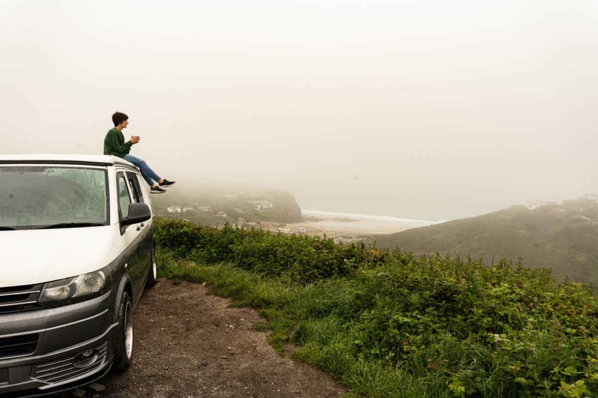 Viaggiatore seduto sul tetto del van con tazza in mano, costa avvolta nella nebbia.