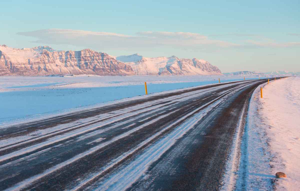 Carreggiata invernale con strisce di ghiaccio e montagne innevate al tramonto.
