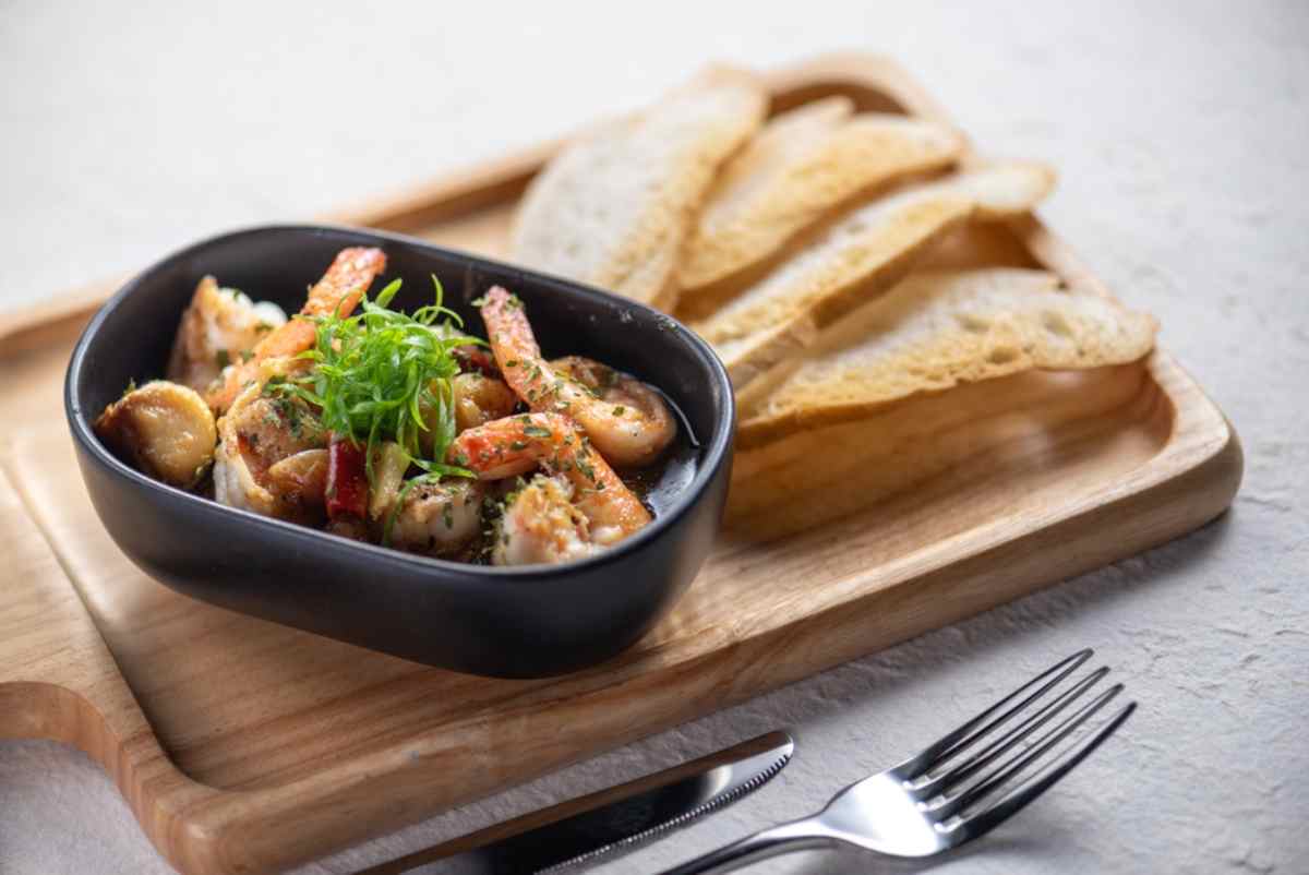 Pirofila con gamberi saltati all&rsquo;aglio, erbe e fette di pane croccante su tagliere.